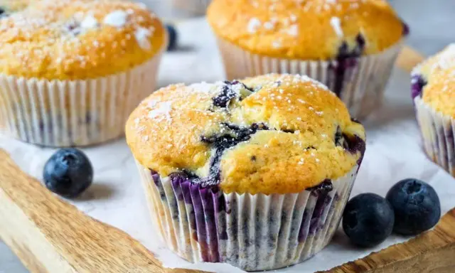 Frisch gebackene Blaubeer Muffins, bestäubt mit Puderzucker, liegen auf einem Holzbrett.