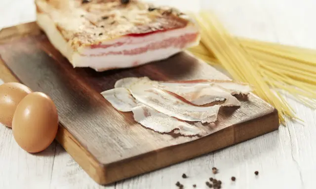 Zutaten für Guanciale Carbonara: ein Stück Guanciale, Eier, Spaghetti und Pfefferkörner auf einem Holzbrett.
