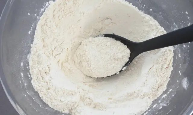 Eine schwarze Löffel hebt eine Portion feines weizenmehl typ 405 aus einer Glasschüssel.