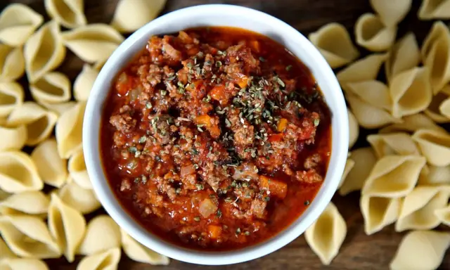 Herzhaftes Ragout alla Bolognese in einer Schüssel, umgeben von ungekochten Muschelnudeln.