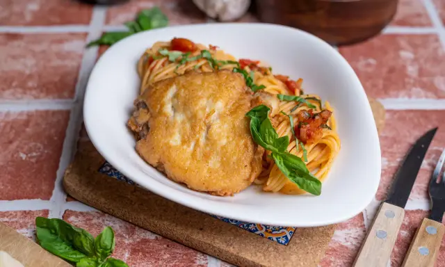 Ein Teller Spaghetti mit Tomatensauce und einem panierten Schnitzel, das ist Piccata. Frisch garniert mit Basilikum.