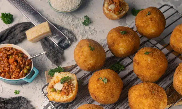 Goldbraune Arancini, gefüllt mit Ragout, liegen auf einem Rost. Zutaten für das Arancini Rezept wie Reis, Parmesan und Hackfleisch sind daneben arrangiert.