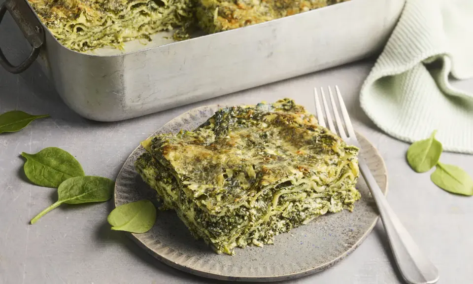 Ein Stück Spinatlasagne auf einem Teller, daneben frische Spinatblätter und eine Gabel. Das Spinatlasagne rezept ist ein Genuss!