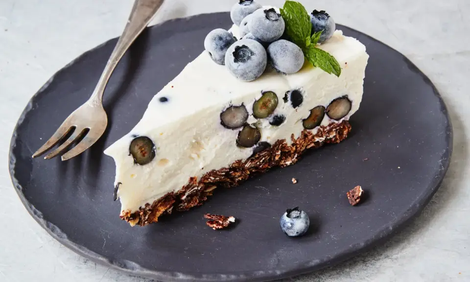 Ein Stück Heidelbeertorte ohne Backen auf schwarzer Platte, garniert mit frischen Heidelbeeren und Minze.