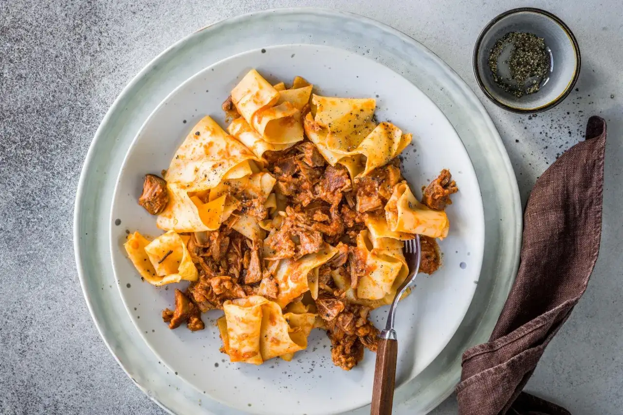 Breite Bandnudeln, was ist Pappardelle, mit herzhaftem Ragout auf einem Teller, garniert mit schwarzem Pfeffer.