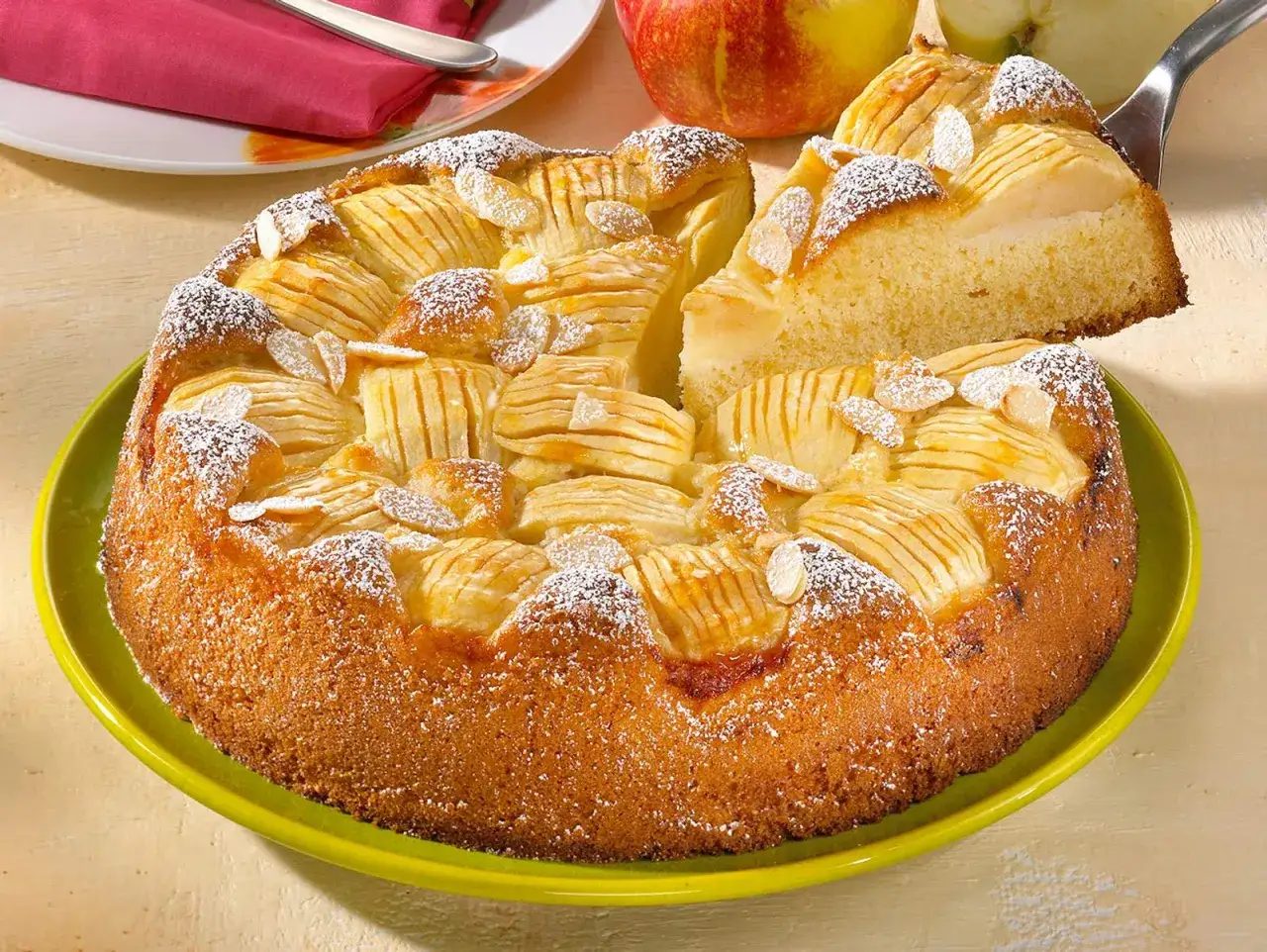 Ein saftiger, versunkener Apfelkuchen nach Omas Rezept, bestreut mit Puderzucker und Mandelblättchen. Ein Stück wird gerade herausgehoben.