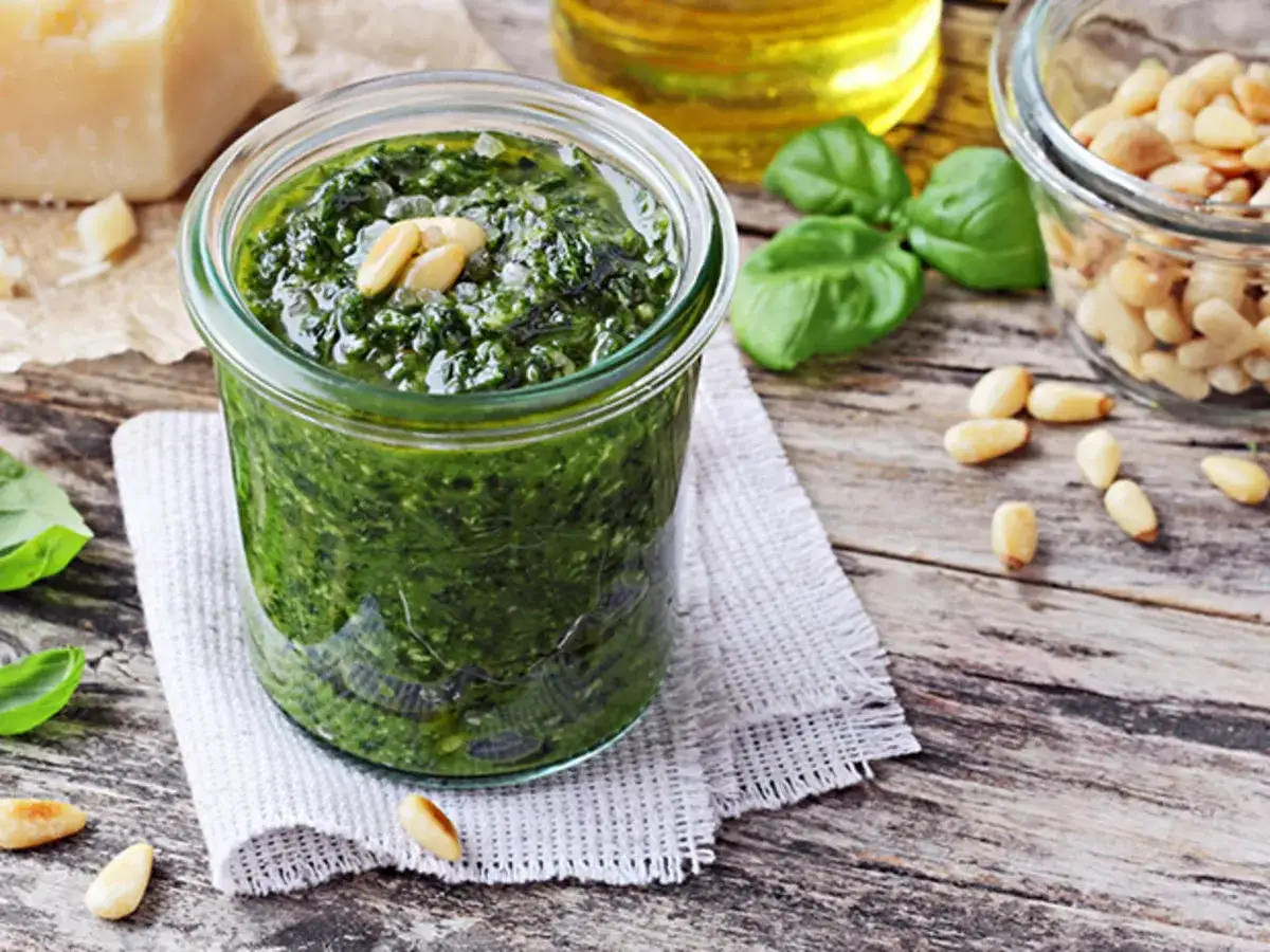 Frisches Pesto-Rezept im Glas, garniert mit Pinienkernen. Daneben Pinienkerne, Basilikum, Parmesan und Olivenöl.
