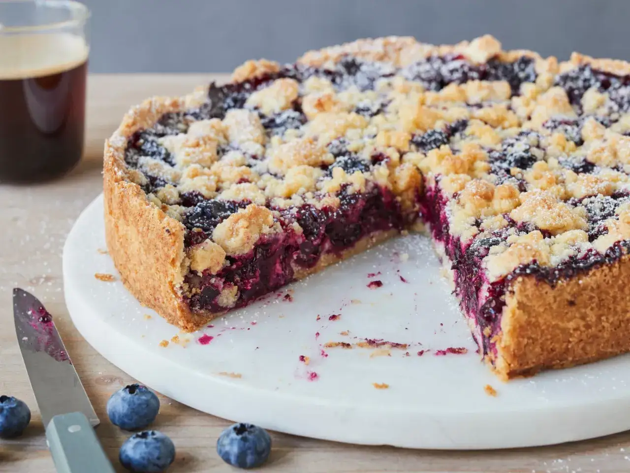 Ein saftiger Heidelbeerkuchen mit Streuseln, bereit zum Genießen. Daneben eine Tasse Kaffee und frische Heidelbeeren.