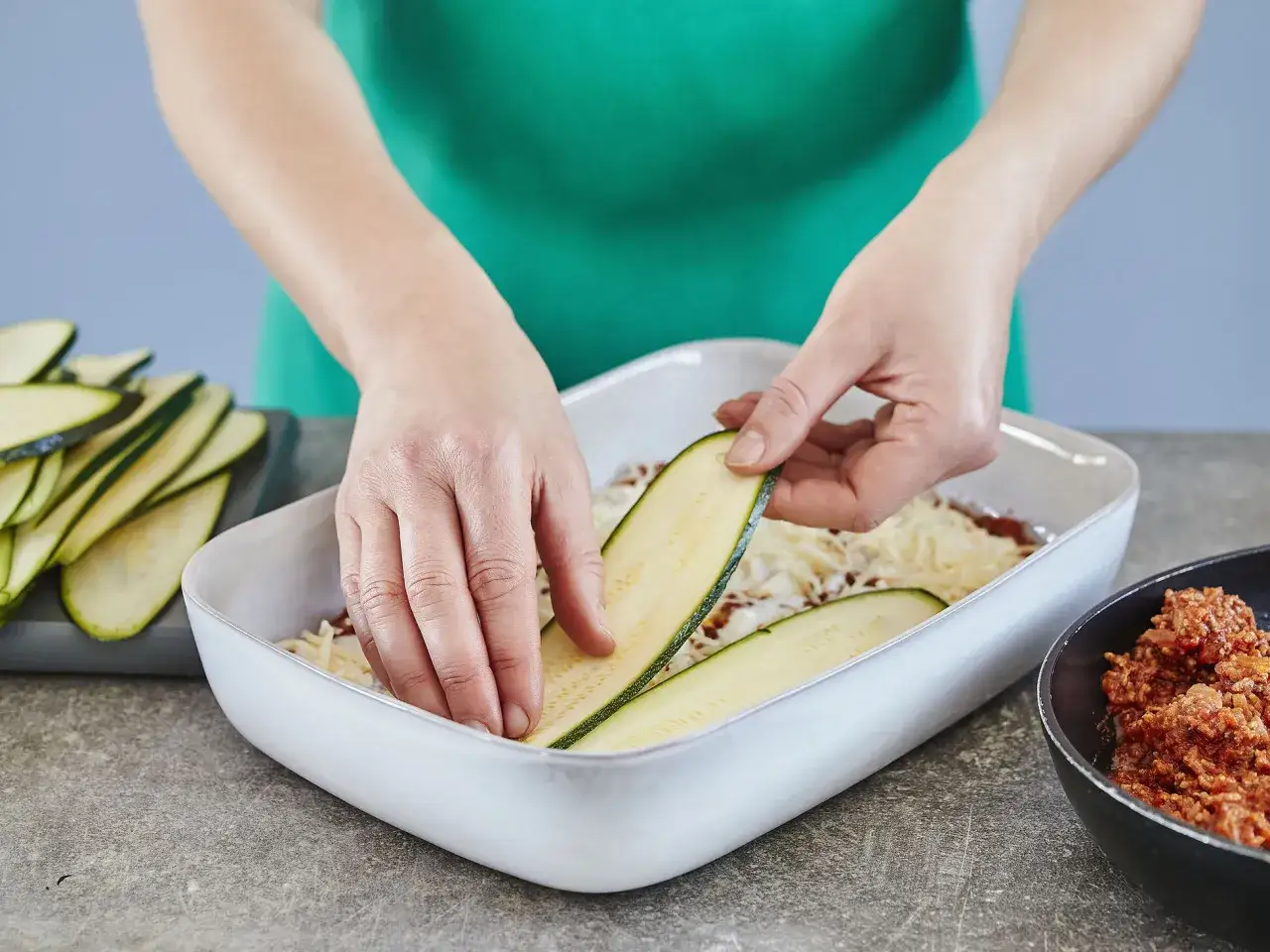 Hände legen Zucchinischeiben auf eine Schicht Käse und Hackfleisch in einer Auflaufform. Die Zubereitung einer gesunden Zucchini-Lasagne beginnt.
