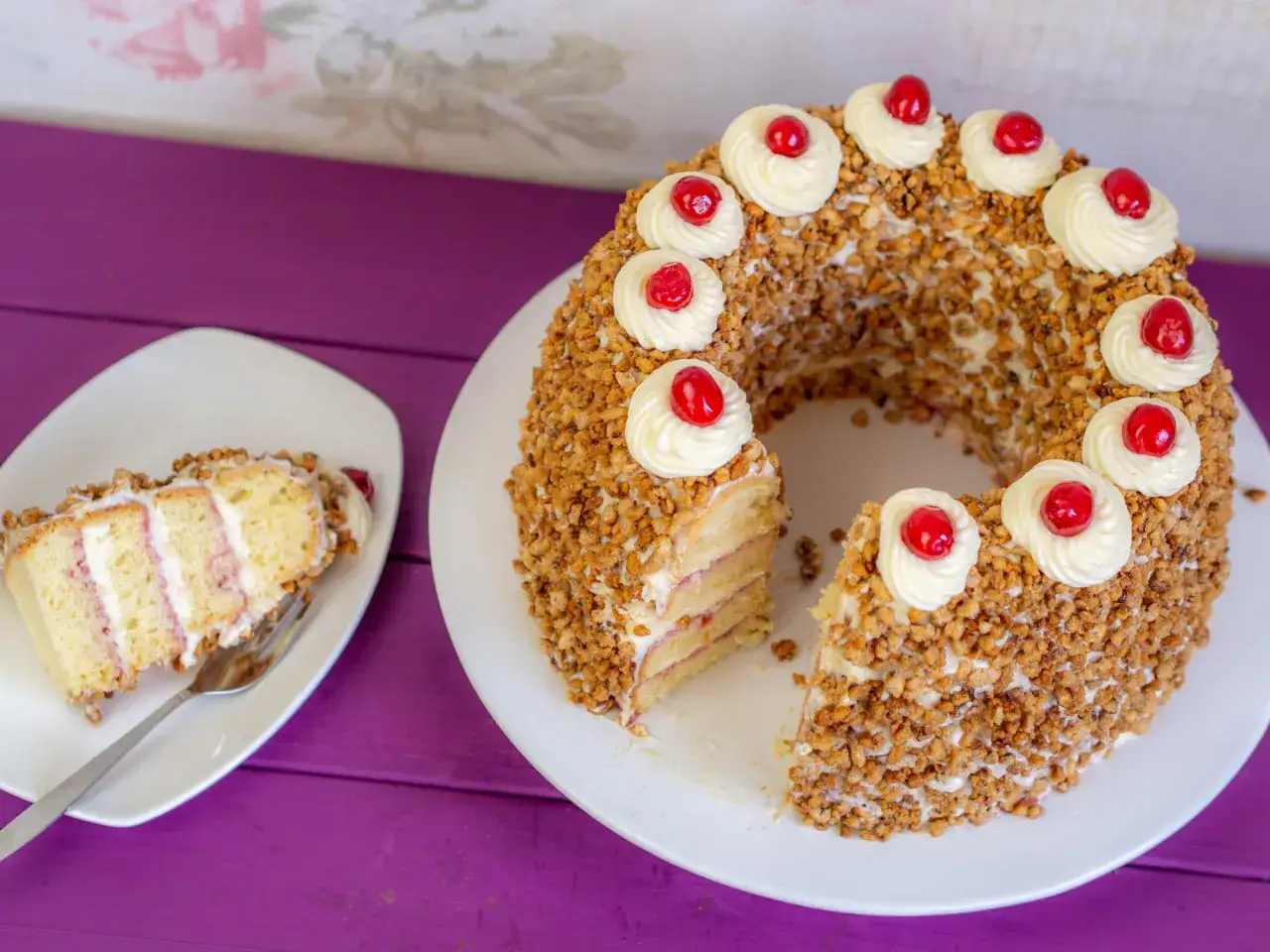 Ein köstlicher Frankfurter Kranz, umhüllt von gehackten Nüssen und verziert mit Sahnetuffs und Kirschen. Ein Stück ist bereits auf einem Teller angerichtet.