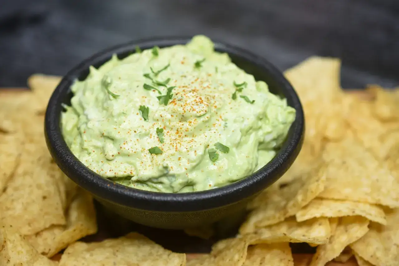 Schüssel mit cremiger Avocado-Creme, garniert mit Petersilie und Paprikapulver, umgeben von Tortilla-Chips.