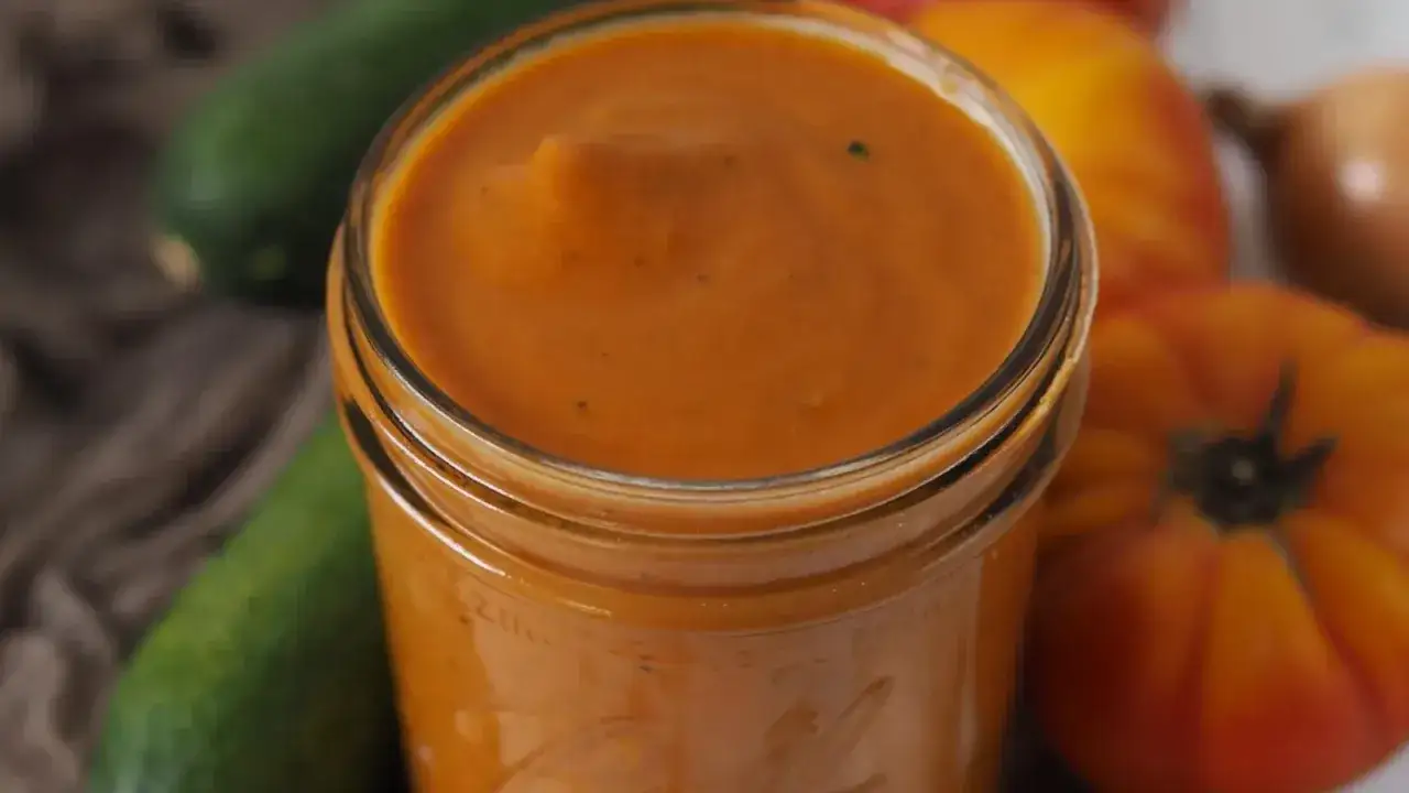 Ein Glas mit leuchtend orangefarbenem Zucchini-Ketchup, umgeben von frischen Zucchini, Tomaten und Zwiebeln.
