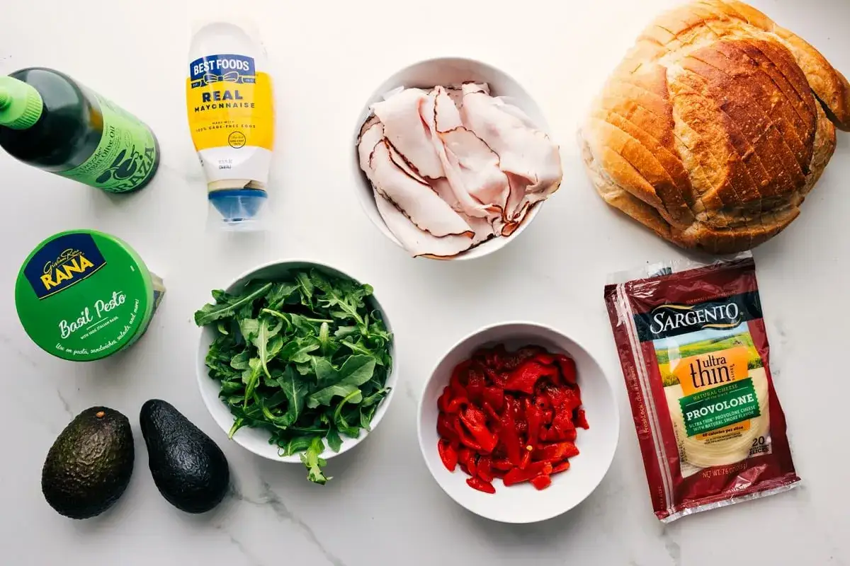 Zutaten f&uuml;r ein leckeres Panini-Rezept: Brot, Putenbrust, Rucola, Paprika, Avocado, Pesto, Mayo und Oliven&ouml;l.
