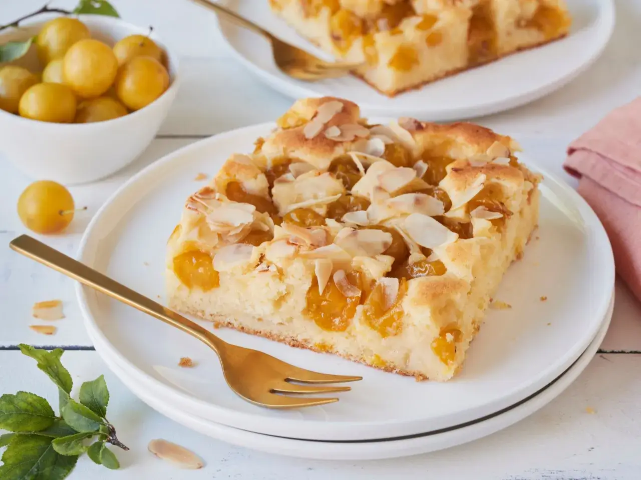Ein Stück saftiger Mirabellenkuchen mit Mandelsplittern auf einem Teller, daneben eine Schale voller frischer Mirabellen.