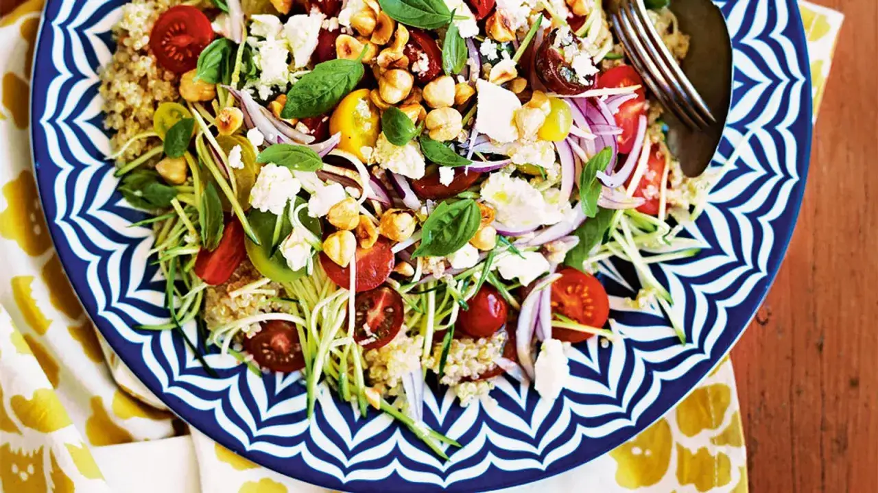 Bunter Quinoa Salat mit Kirschtomaten, Zucchini-Spaghetti, Feta und N&uuml;ssen auf einem blau-wei&szlig;en Teller.