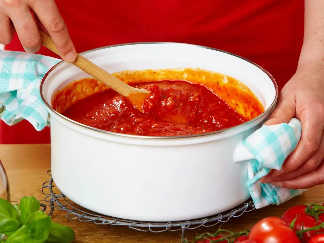 H&auml;nde r&uuml;hren in einem Topf mit k&ouml;stlicher, selbstgemachter Tomatensauce. Frische Tomaten und Basilikum liegen daneben.