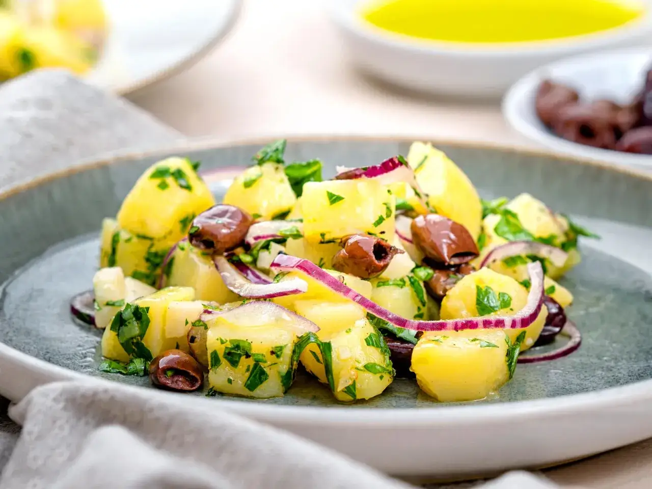 Ein k&ouml;stlicher italienischer Kartoffelsalat mit Oliven, roten Zwiebeln und frischer Petersilie auf einem grauen Teller.