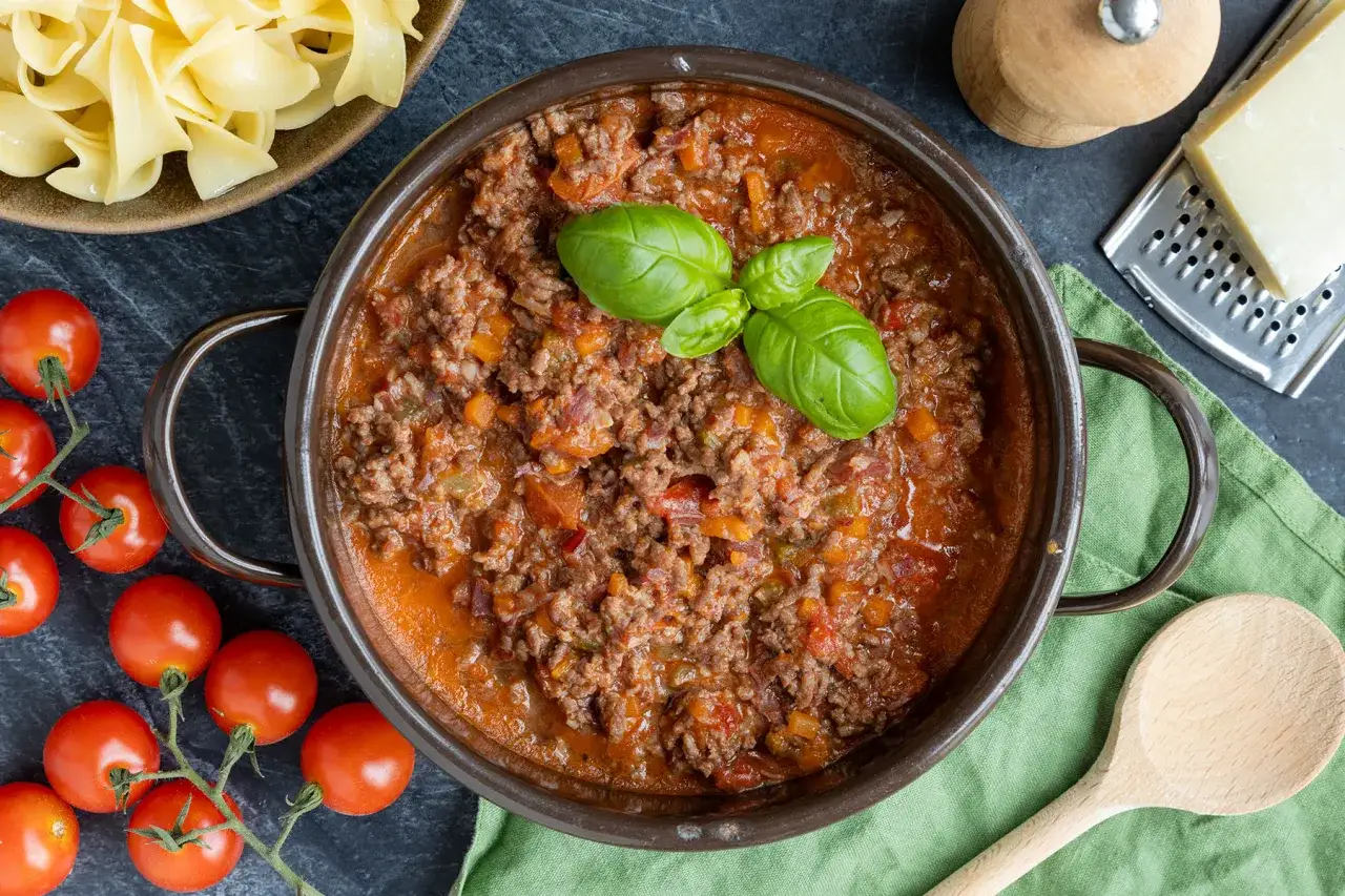 Herzhaftes Ragout alla Bolognese in einem Topf, garniert mit Basilikum. Daneben frische Tomaten, Bandnudeln und Parmesan.