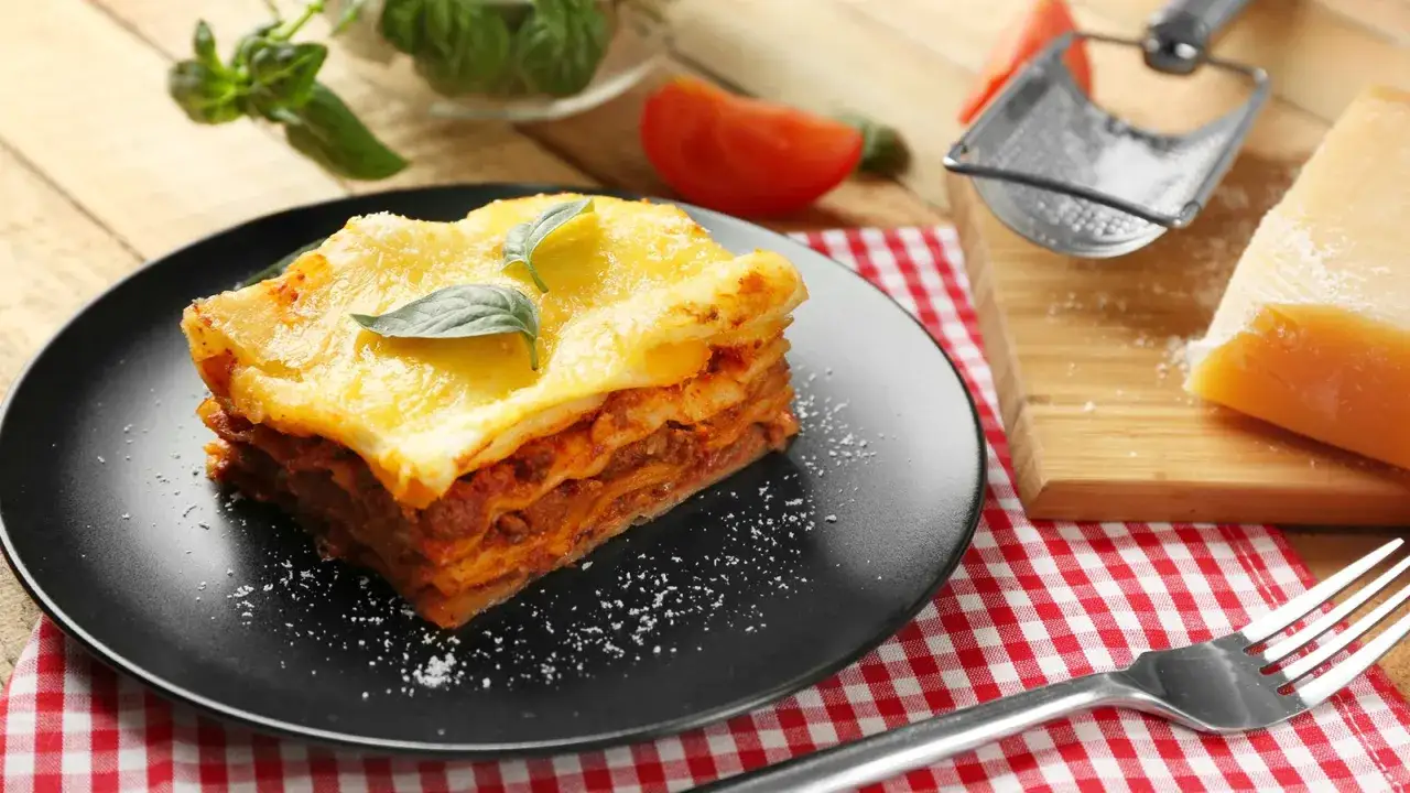 Ein Stück Lasagne mit reichlich Bechamelsauce und Käse überbacken, garniert mit Basilikum. Daneben ein Stück Parmesan und eine Gabel.