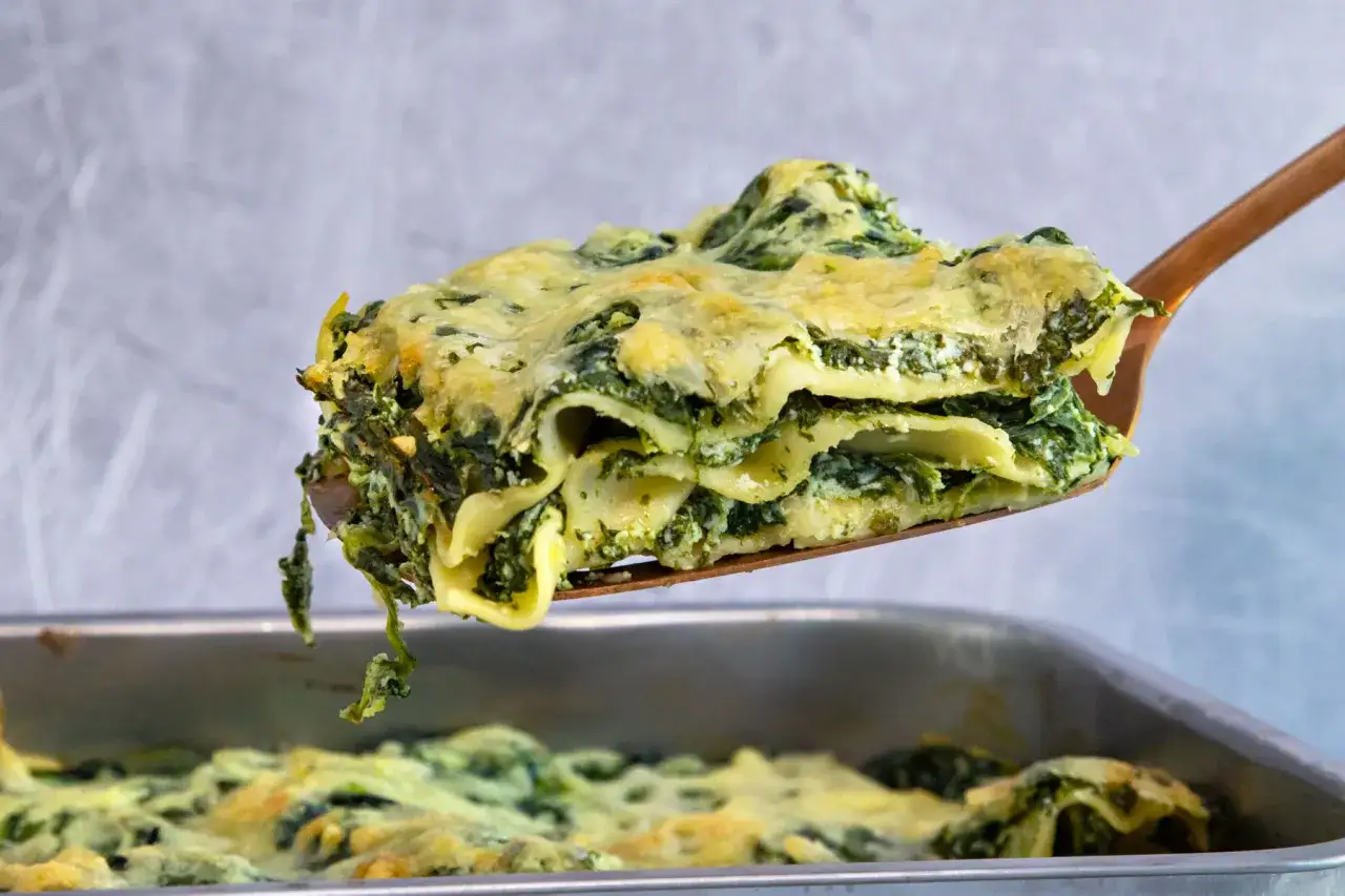 Ein St&uuml;ck Spinatlasagne wird aus einer Auflaufform gehoben. Das Gericht ist mit K&auml;se &uuml;berbacken und sieht k&ouml;stlich aus.