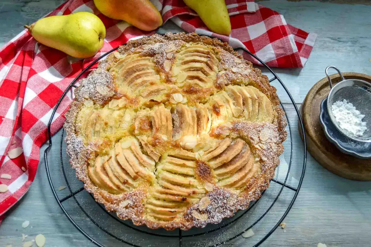 Eine k&ouml;stliche Birnen Tarte mit Mandelsplittern und Puderzucker best&auml;ubt, daneben frische Birnen und ein kariertes Tuch.