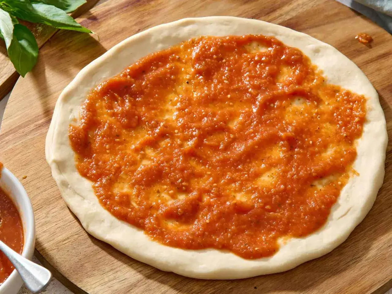 Frische Pizza mit selbstgemachter pizza sauce wie in der pizzeria, bereit für den Ofen. Ein Hauch von Basilikum liegt daneben.
