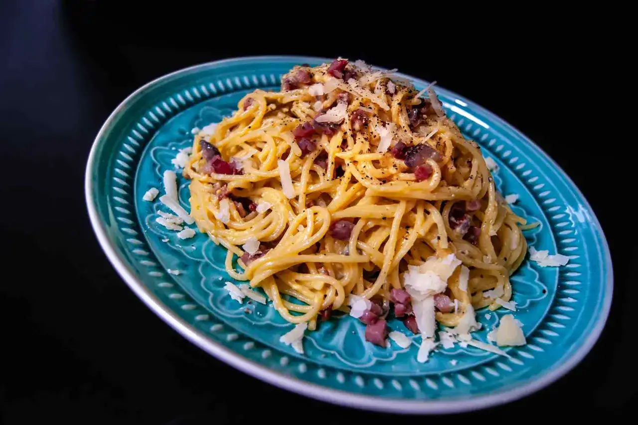 Ein Teller Spaghetti Carbonara mit Guanciale, Käse und schwarzem Pfeffer auf einem türkisfarbenen Teller.