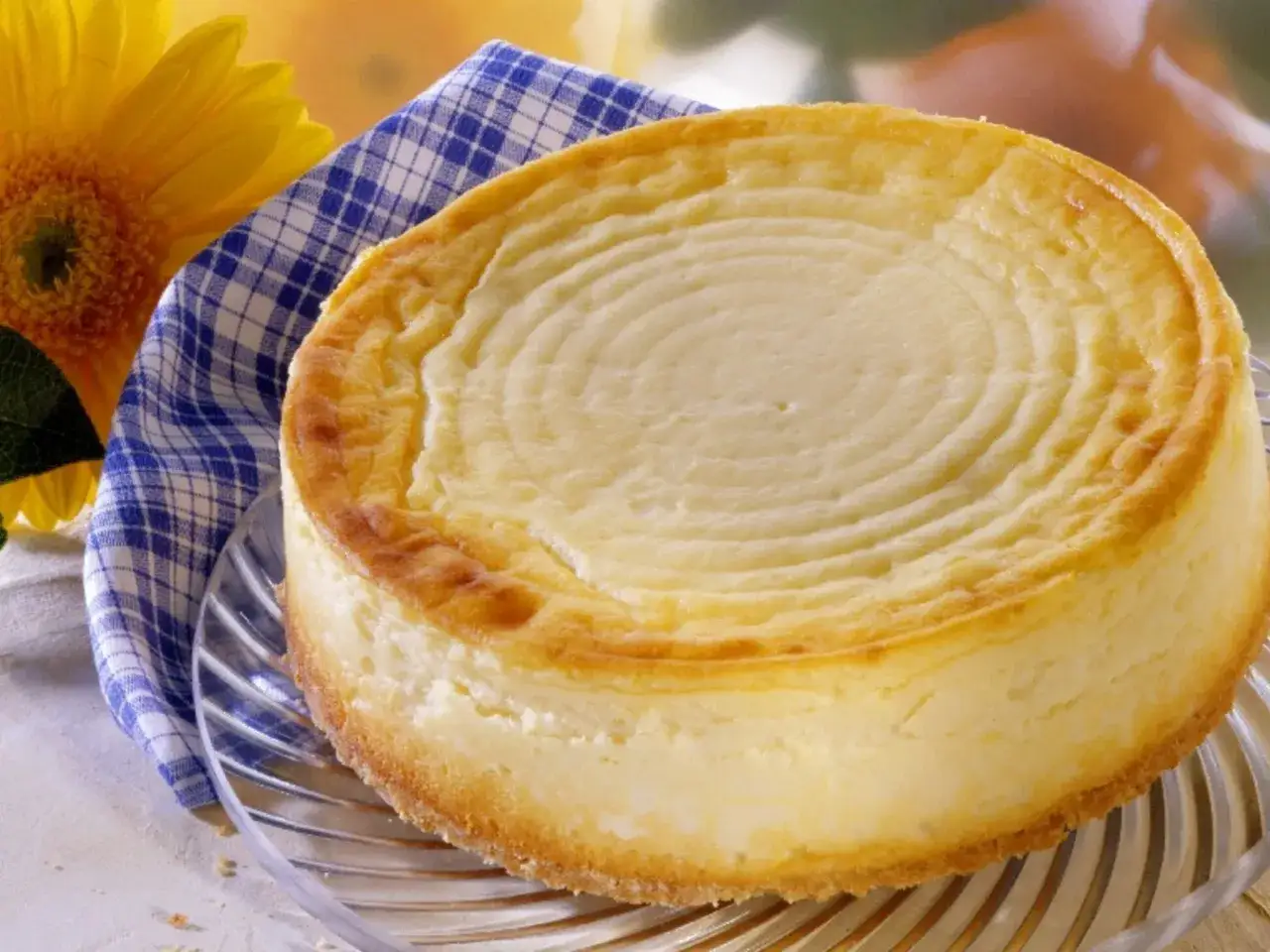 Ein goldbrauner, cremiger american cheesecake mit Ringen auf der Oberfl&auml;che, neben einer gelben Blume und einem blau-wei&szlig; karierten Tuch.