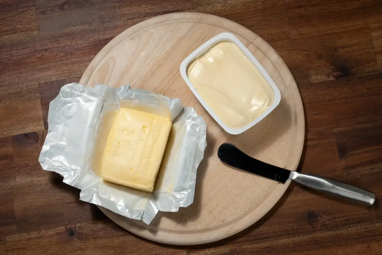 Ein Block Butter in Folie und eine Schale mit Butter oder Margarine liegen auf einem Holzbrett. Ein Messer liegt daneben.