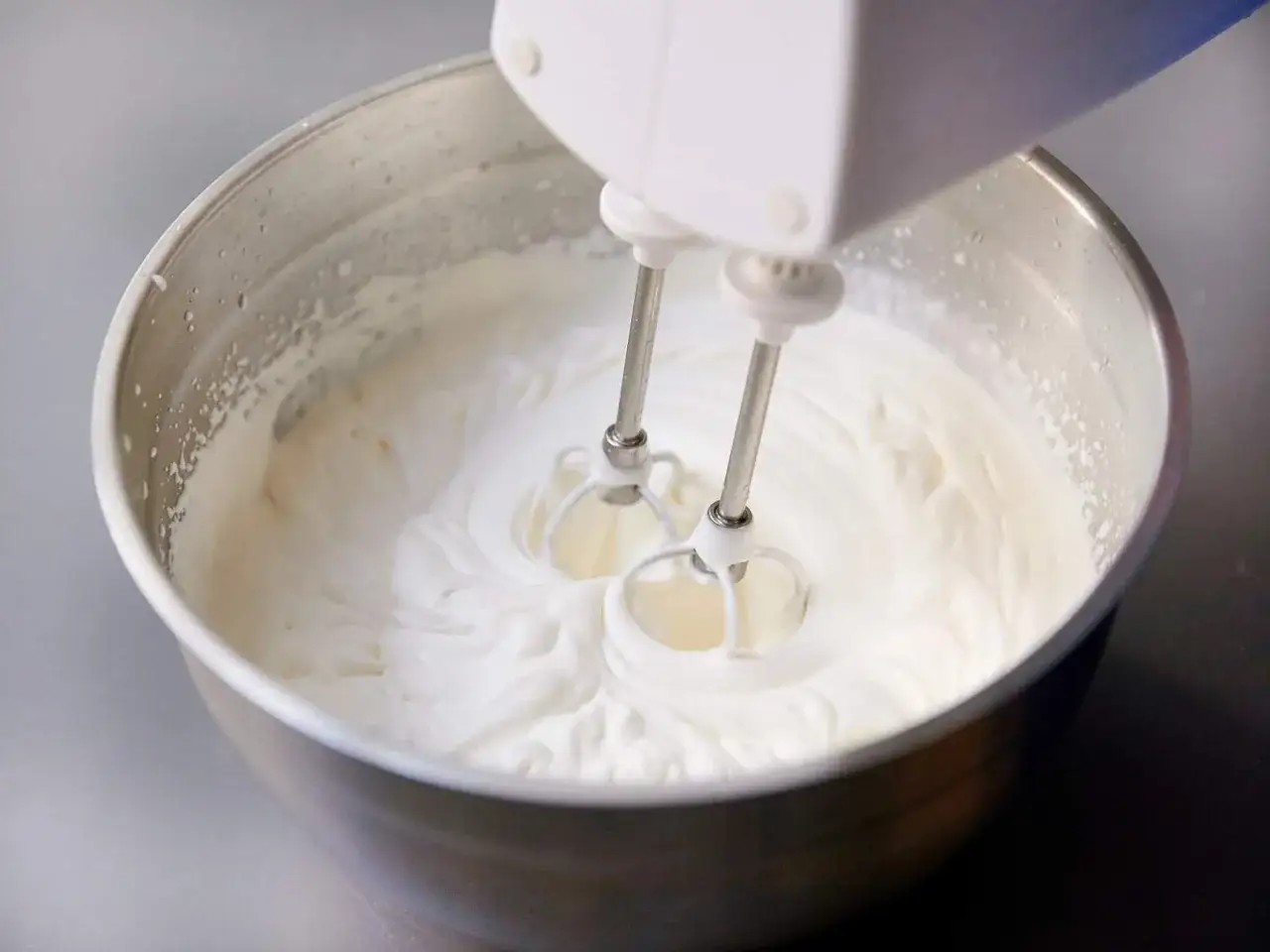 Ein Handmixer schl&auml;gt gerade Sahne in einer Edelstahlsch&uuml;ssel. Die wei&szlig;e, fluffige Masse wirbelt um die Quirlen.