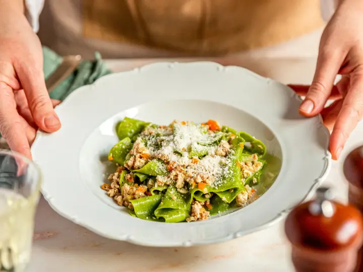 Grüne Bandnudeln mit herzhaftem ragu bianco und Parmesan bestreut, serviert auf einem weißen Teller.